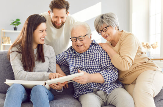 Elderly couple and grown son and daughter sitting together on the couch at home, talking, laughing and looking through a family photo album. Happy old parents share memories with adult children.