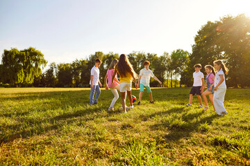 Obraz premium Happy children boys and girls running towards ball playing together in the park on the green grass on a summer holidays. Teenage laughing kids in casual clothes playing football in the garden.