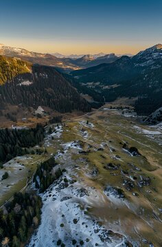 Captivating aerial snapshot of a snowy alpine landscape blending green meadows and dense forests with majestic mountains under a golden hour sky, shot on a Sony Alpha A7 III.