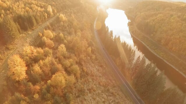FPV drone: Dramatic aerial shot. Cars on scenic road by water dam. Sun flares through vibrant fall foliage. Perfect for car ads or commercials, travel and road trip docs. LuPa Creative