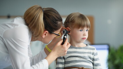 Female pediatrician looking at ear of little girl using otoscope in clinic. Hearing loss diagnosis...