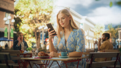 Attractive Female Sitting Alone in a Street Cafe, Having a Cup of Coffee and Croissant. Young Woman Using Smartphone, Chatting with Friends Online, Checking Social Media and Work Emails