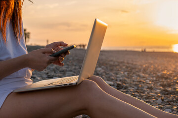 Close up of woman working with laptop and mobile phone on the beach by the sea during sunset....