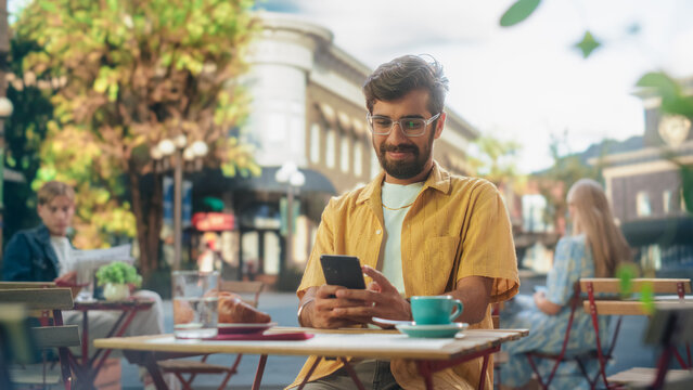 Handsome Multiethnic Man Sitting on a Terrace in a Cafe, Having a Cup of Coffee with Pastry. Happy Indian Man Connecting with Friends Online, Replying to Social Media Posts and Emails on a Smartphone - Powered by Adobe
