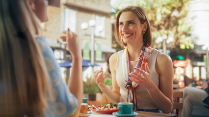 Two Beautiful Female Friends Sitting in Front of Each Other in an Outdoors Restaurant, Eating Healthy Vegetarian Salad and a Dessert. Girls Excited to Meet and Talk About Their Summer Holiday Plans