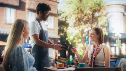 Two Female Friends Ordering Delicious Cake Desserts From a Member of the Staff in a Cafe. Handsome Multiethnic Waiter Holding a Terminal, Female Using Her Smartphone for a Contactless NFC Payment