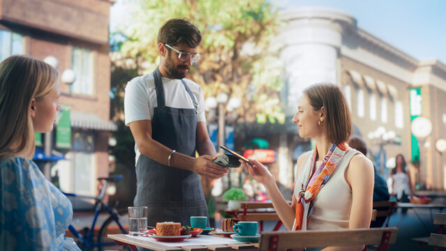 Female is Making a Payment to a Waiter with Her Smartphone. Friends are Enjoying Cake Desserts on a Terrace in a Cafe and Closing the Bill with a Contactless NFC Function on a Mobile Device