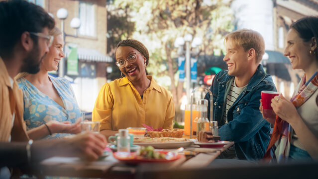Diverse Group of Friends Enjoying Leisure Time in a Street Cafe. Young Women and Men Sitting Behind a Table, Having Fun and Joyful Conversations. Black African Girl Sharing Motivational Work Stories
