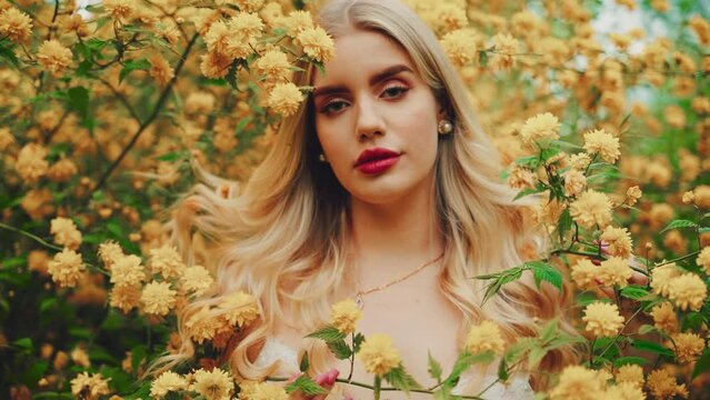 Woman beauty face close-up eyes lips in yellow flowers. sexy girl fashion model looking camera. spring garden tree enjoy smell. Blonde curly hair lady inhales floral scent white dress boho style. 4k