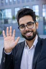 Vertical close-up photo of a young smiling Indian man in a suit and glasses talking to the camera on a video call, waving and greeting.