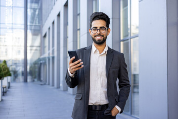 Fototapeta premium Portrait of a smiling young Indian businessman in a suit standing outside an office building, holding his hand in his pocket, using the phone and looking at the camera.