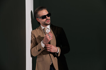 unshaved cool man in brown suit fixing bracelets and looking to side