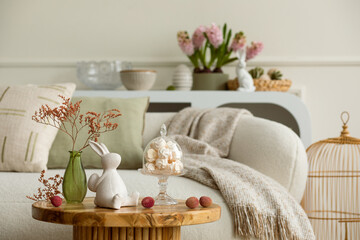 Warm and cozy composition of easter living room interior with coffee table, white sofa, colorful easter eggs, easter bunny, hyacinth, vase with branch and personal accessories. Home decor. Template.