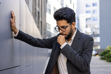 A young Indian man in a business suit is standing near the office, leaning his hand on the wall and suffering from a strong cough, covering his mouth with his hand.
