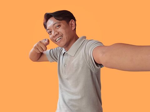 Handsome young man wearing gray t-shirt standing isolated over orange background, taking a selfie, pointing finger at camera