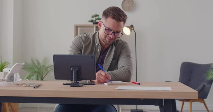 Young man studying at home. Happy, smiling male student in eyeglasses sitting at table with modern tablet computer, attending online training course lesson, writing notes, typing on mini keyboard 