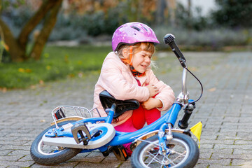 Cute little girl sitting on the ground after falling off her bike. Upset crying preschool child with safe helmet getting hurt while riding a bicycle. Active family leisure with kids.