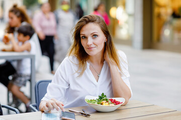 Young beautiful woman eating fresh salad in outdoor restaurant. Woman enjoying lunch bowl with fresh vegetables. Healthy food. On summer day.