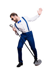 Full size fullbody portrait of famous creative singer in blue pants with suspenders, black glasses, singing hit with open mouth in mic gesture with hand isolated on grey background