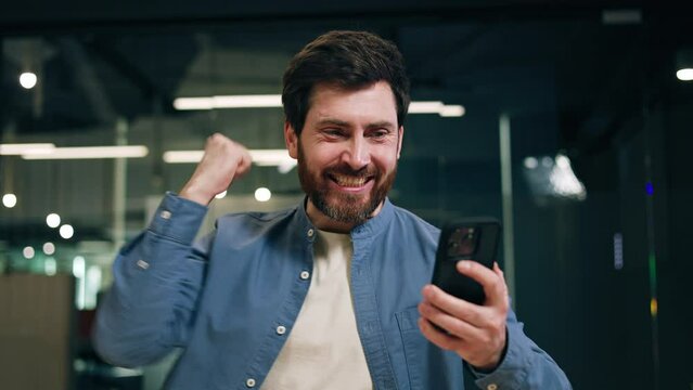 Amazed guy in denim shirt celebrating success while winning financial prize for achievement in mobile phone application. Surprised digital device user reaching goal and getting massive reward.
