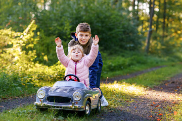 Two happy children playing with big old toy car in autumn forest, outdoors. Kid boy pushing and driving car with little toddler girl, cute sister inside. Laughing and smiling kids. Lovely family