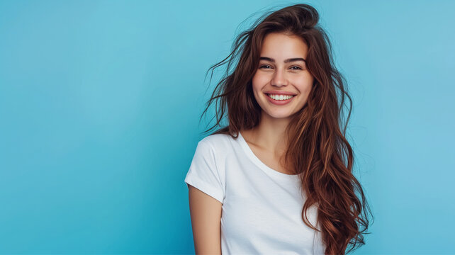 Pretty young smiling woman with dark hair on a blue background with copy space. Cute girl in a simple white T-shirt.