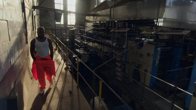 Slowmo portrait of Black male worker wearing orange coverall and white undershirt strolling along dark industrial factory then posing for camera