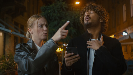 Indian Arabian Latino man and Caucasian woman arguing conflict using mobile phone telephone walking city street evening outdoors choice couple annoyed argument disagree displeased quarrel relationship