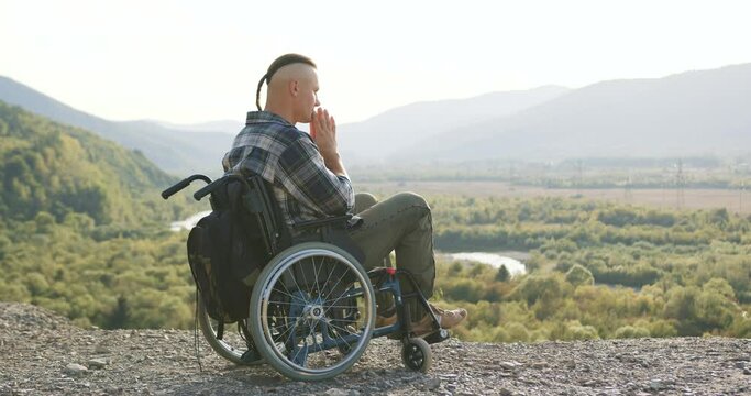 Concentrated man with little ponytail which sitting in wheelchair on the hill and looking around on the mount