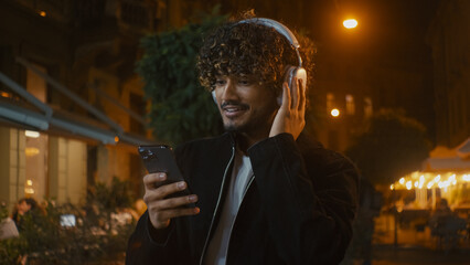Indian Latino Arabian happy man walking city street outdoors evening listening music headphones earphones using smartphone mobile phone gadget handsome guy stylish male smile satisfied weekend modern
