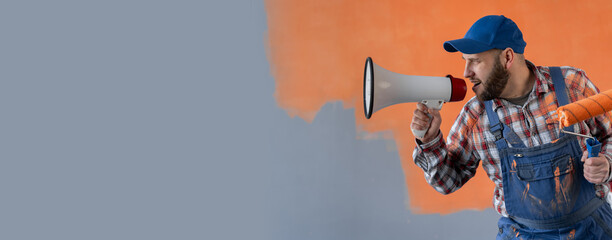 Handsome bearded painter with painting roller shouting loud by megaphone standing near wall in...