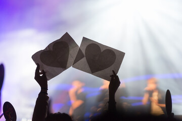 Anonymous silhouetted hands holding heart-shaped cutouts at a lively concert, capturing a moment of unity and fan adoration in a glowing stage ambiance