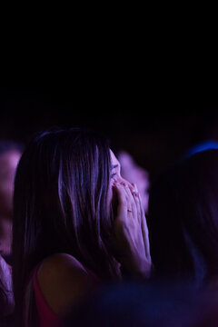A woman in a crowd at a concert is captured in a profound moment of emotion, her hands covering her face against a backdrop of ambient stage lighting