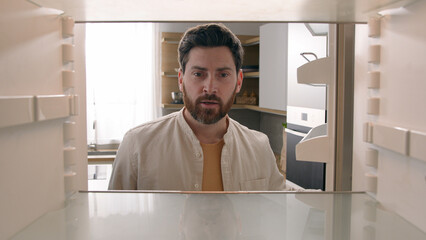 Hungry Caucasian bachelor man looking for food in empty fridge finding no meal products feeling sad...