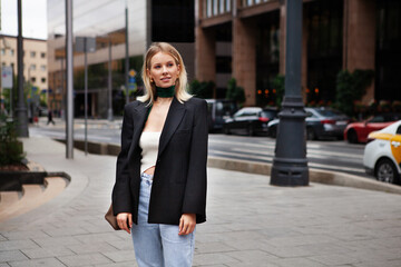 Fototapeta premium Beautiful young stylish blonde woman on city street against backdrop of building, wearing fashionable clothes, black blazer and silk scarf. Fashion model looks away