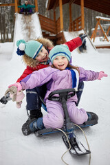 Laughing little girl and boy sit on sled after descent from wooden slide covered with snow on winter day