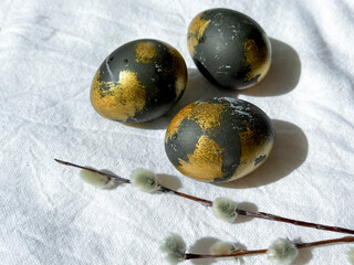 Dark blue and gold Easter eggs lie on a white light tablecloth under the rays of the sun, willow branches lie nearby. An Easter card