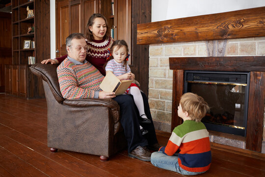Father reads book aloud sitting in armchair near fireplace, mother and two children liten to him