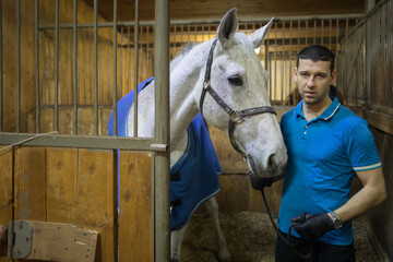 Handsome man in gloves and white horse in harness are in stable