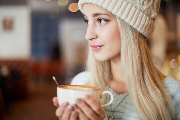 Young blonde woman in knitted hat holds cup of coffee in hands