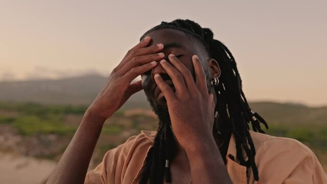 Closeup of a serene man with dreadlocks meditating at sunset, touching his face in a moment of self-reflection and peace.