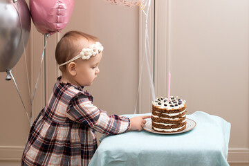  ute little girl blows out candles on a birthday cake at against backdrop of balloons. Child's birthday. Little girl in blowing out candles on birthday chocolate cake on her birthday