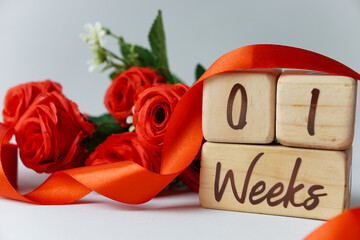 1 week gestational age milestone written on a wooden cube with red roses and ribbons, and a white...