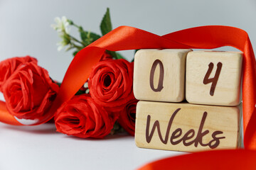 4 week gestational age milestone written on a wooden cube with red roses and ribbons, and a white...