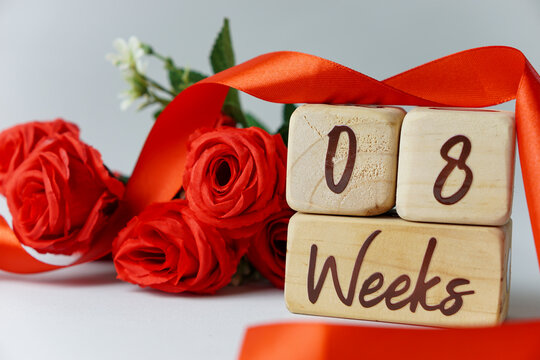 8 week gestational age milestone written on a wooden cube with red roses and ribbons, and a white background