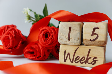 15 week gestational age milestone written on a wooden cube with red roses and ribbons, and a white...