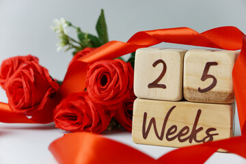25 week gestational age milestone written on a wooden cube with red roses and ribbons, and a white...