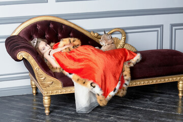 Girl with crown on head lies on couch covered by red cloak, lynx cub sits near