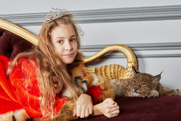 Girl in red cloak with crown on head lies on couch with lynx cub