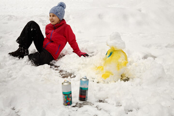 Smiling girl sits on snow near head of snowman painted by spray